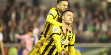 Η ΑΕΚ νίκησε τον Παναθηναϊκό, το βράδυ της Κυριακής (30/11), με 2-3 χάρη σε ένα πέναλτι που εκτέλεσε ο Γιόβιτς, στις καθυστερήσεις της αναμέτρησης που έγινε στη Λ. Αλεξάνδρας, στο πλαίσιο της 12ης αγωνιστικής της Super League. Το newsletter του iEidiseis καθημερινά στο inbox σου. Κάνε εγγραφή εδώ. Συγκεκριμένα, σε ένα από τα πιο εντυπωσιακά ντέρμπι της σεζόν, το ματς είχε τα πάντα – δύο αποβολές, ανατροπές στο σκορ, ένταση και ένα φινάλε που «έσπασε καρδιές». Ο αγώνας Παναθηναϊκός – ΑΕΚ Ο Παναθηναϊκός μπήκε στο παιχνίδι δίνοντας χώρο στην ΑΕΚ, με την κατοχή των «κιτρινόμαυρων» να αγγίζει μέχρι και το 67%. Οι φιλοξενούμενοι αναζητούσαν την κάθετη πάσα, και τελικά στο 17΄ βρήκαν τον δρόμο προς τα δίχτυα. Ήταν μία εκτέλεση φάουλ-κομπίνα από τον Μάνταλο, ο οποίος έδωσε πάσα στον Ρότα. Ο δεξιός μπακ σέντραρε στο δεύτερο δοκάρι και ο Γιόβιτς πήρε την κεφαλιά για το 0-1. Όπως φάνηκε, η φάση ήταν… δουλεμένη. Στο 28′, ο Παναθηναϊκός είχε την ευκαιρία να ισοφαρίσει, όταν μέσω VAR καταλογίστηκε πέναλτι σε μαρκάρισμα του Πήλιου στον Τετέ. Ωστόσο, ο Στρακόσια «νίκησε» τον Κάρολ Σφιντέρσκι από τα 11 βήματα, κρατώντας ανέπαφη την εστία του. Η ΑΕΚ «χτύπησε» ξανά πριν από το ημίχρονο. Στο 45+2′ ο Τουμπά ανέτρεψε τον Γιόβιτς, ο οποίος εκτέλεσε το πέναλτι α λα Πανένκα για το 0-2. Οι «πράσινοι» είχαν προηγουμένως διαμαρτυρηθεί για χέρι του Ρέλβας. Το παιχνίδι συνεχίστηκε σε χαμηλό τέμπο, καθώς η ΑΕΚ διαχειριζόταν το προβάδισμά της. Ο Παναθηναϊκός άλλαξε σχηματισμό στο 62′ (4-2-3-1), όμως στο 64′ έμεινε με 10 παίκτες. Ύστερα από λάθος του Πάλμερ Μπράουν, ο Τουμπά ανέτρεψε τον Γκατσίνοβιτς που έβγαινε τετ-α-τετ και αποβλήθηκε. Παρ’ όλα αυτά, οι «πράσινοι» όχι μόνο δεν κατέρρευσαν, αλλά παρουσιάστηκαν πιο απειλητικοί. Στο 71′ ο Ζαρουρί σέντραρε και ο Τετέ με κεφαλιά έκανε το 1-2, εκμεταλλευόμενος το γλίστρημα του Στρακόσια. Πέντε λεπτά αργότερα, ο Τσέριν άγγιξε την ισοφάριση. Στο 85′ αποβλήθηκε και ο Μάριν, αποκαθιστώντας την αριθμητική ισορροπία. Ο Παναθηναϊκός έριξε στο γήπεδο τον Ντέσερς και επιτέθηκε μαζικά για την ισοφάριση. Έπειτα, στο 90′ ο Παναθηναϊκός ολοκλήρωσε την ανατροπή της εικόνας του αγώνα. Ο Τζούρισιτς με ένα εντυπωσιακό αριστερό σουτ «έγραψε» το 2-2, ξεσηκώνοντας τη Λεωφόρο. Ωστόσο, στο 90+3′, ύστερα από σύγχυση στην περιοχή των γηπεδούχων, ο Κοϊτά ανατράπηκε από τον Ντραγκόφσκι. Ο Γιόβιτς πήρε ξανά την ευθύνη από τα 11 μέτρα και ευστόχησε, διαμορφώνοντας το 2-3 και ολοκληρώνοντας το χατ-τρικ του. Στα εναπομείναντα λεπτά, η ΑΕΚ άγγιξε και το 2-4 με τον Γιόβιτς, αλλά ο Κώτσιρας έσωσε πάνω στη γραμμή. Το σκορ δεν άλλαξε.